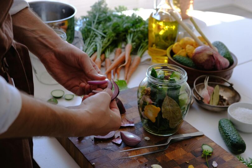 Close-up of hands preparing pickled vegetables in a jar with fresh carrots and cucumbers.